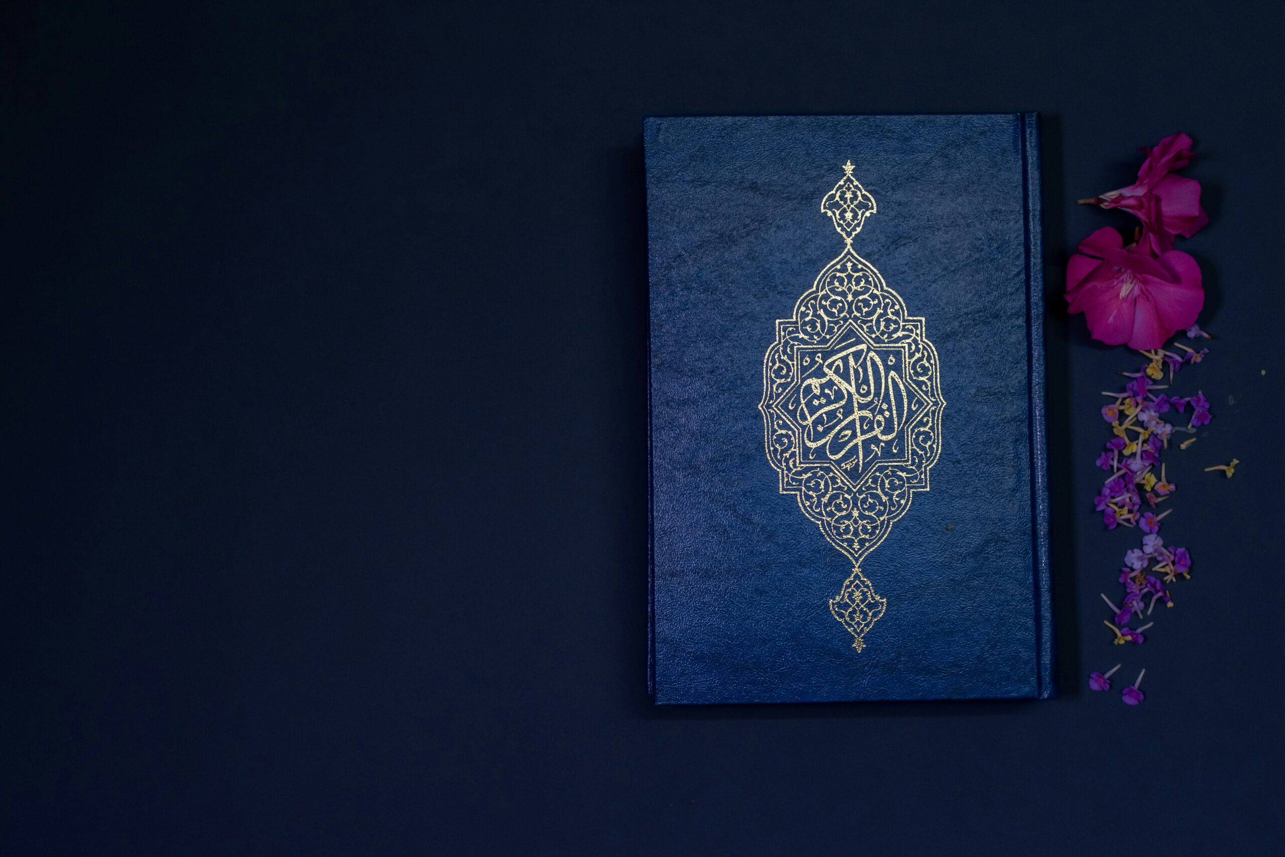 Blue Quran next to a red flower on a blue table.