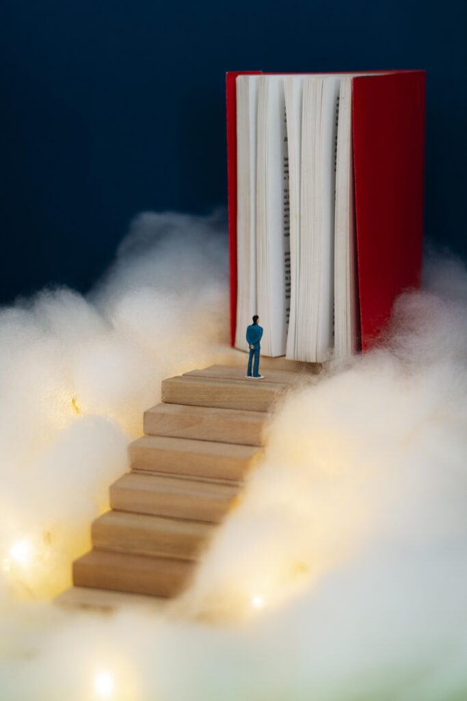 A miniature figure stands on wooden steps leading to a giant open book, surrounded by soft clouds and twinkling lights.