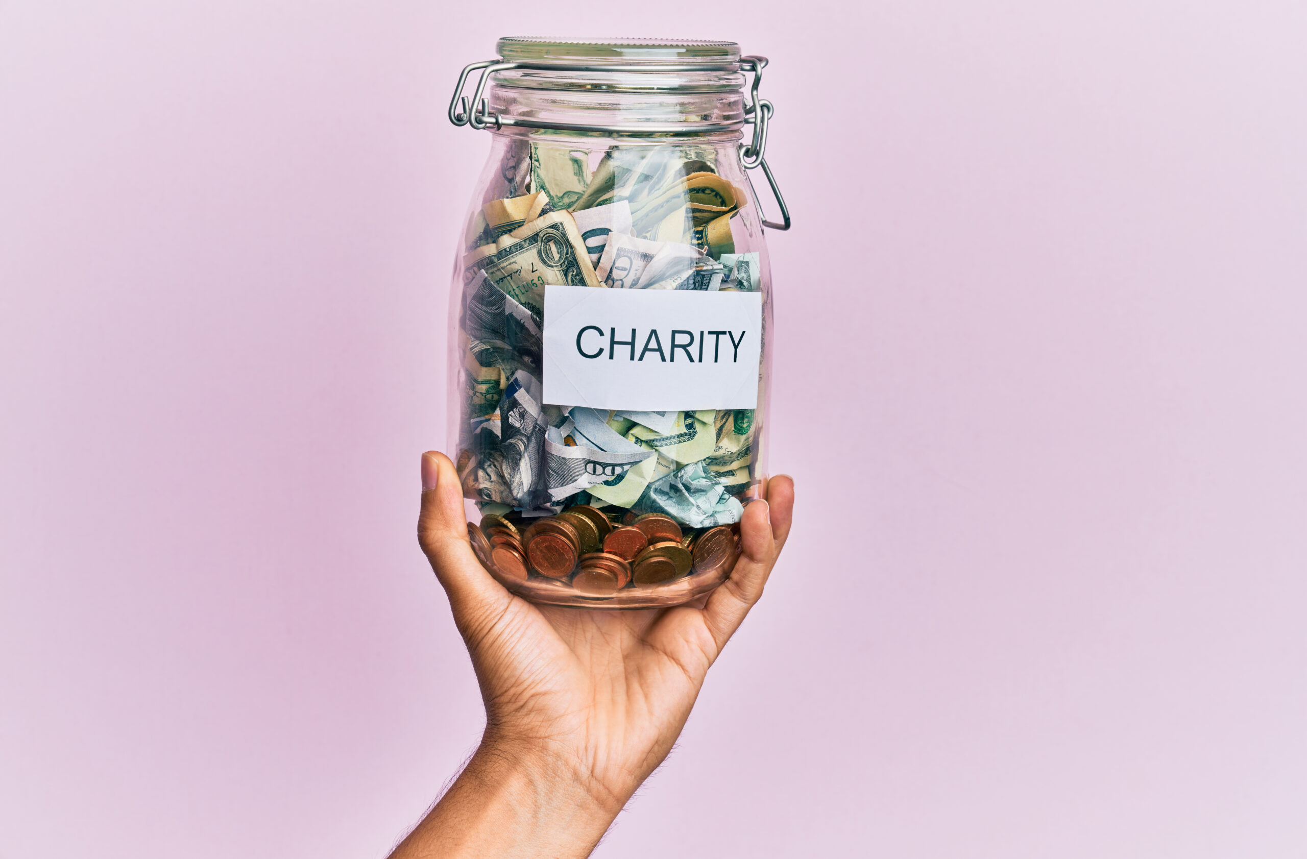 The hand of a Muslim man holding a charity jar with dollars over an isolated pink background.