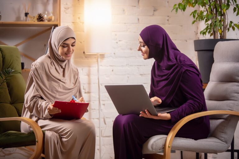 Two women in hijabs share a friendly discussion, one taking notes on a clipboard while the other works on a laptop, in a cozy, well-lit space.