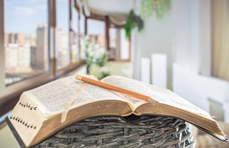 An open book rests on a woven basket, with a pencil placed on top, inviting reflection and study. In the background, large windows showcase a cityscape, adding a modern and airy ambiance to the scene.