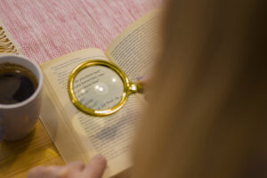 A close-up of a book opened to a page with text, where a magnifying glass rests on the words, accompanied by a cup of coffee on a cozy table setting.
