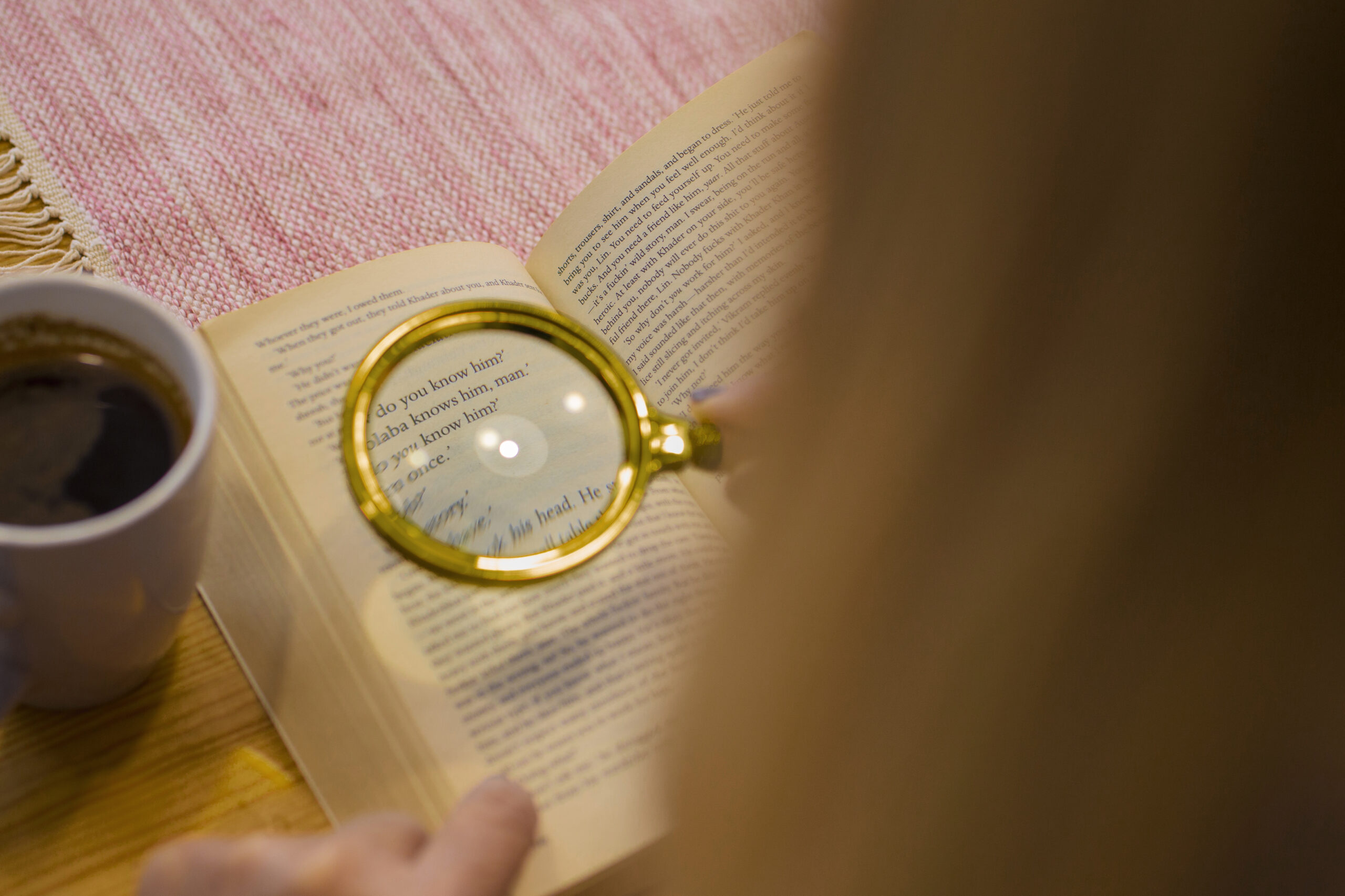 A close-up of a book opened to a page with text, where a magnifying glass rests on the words, accompanied by a cup of coffee on a cozy table setting.