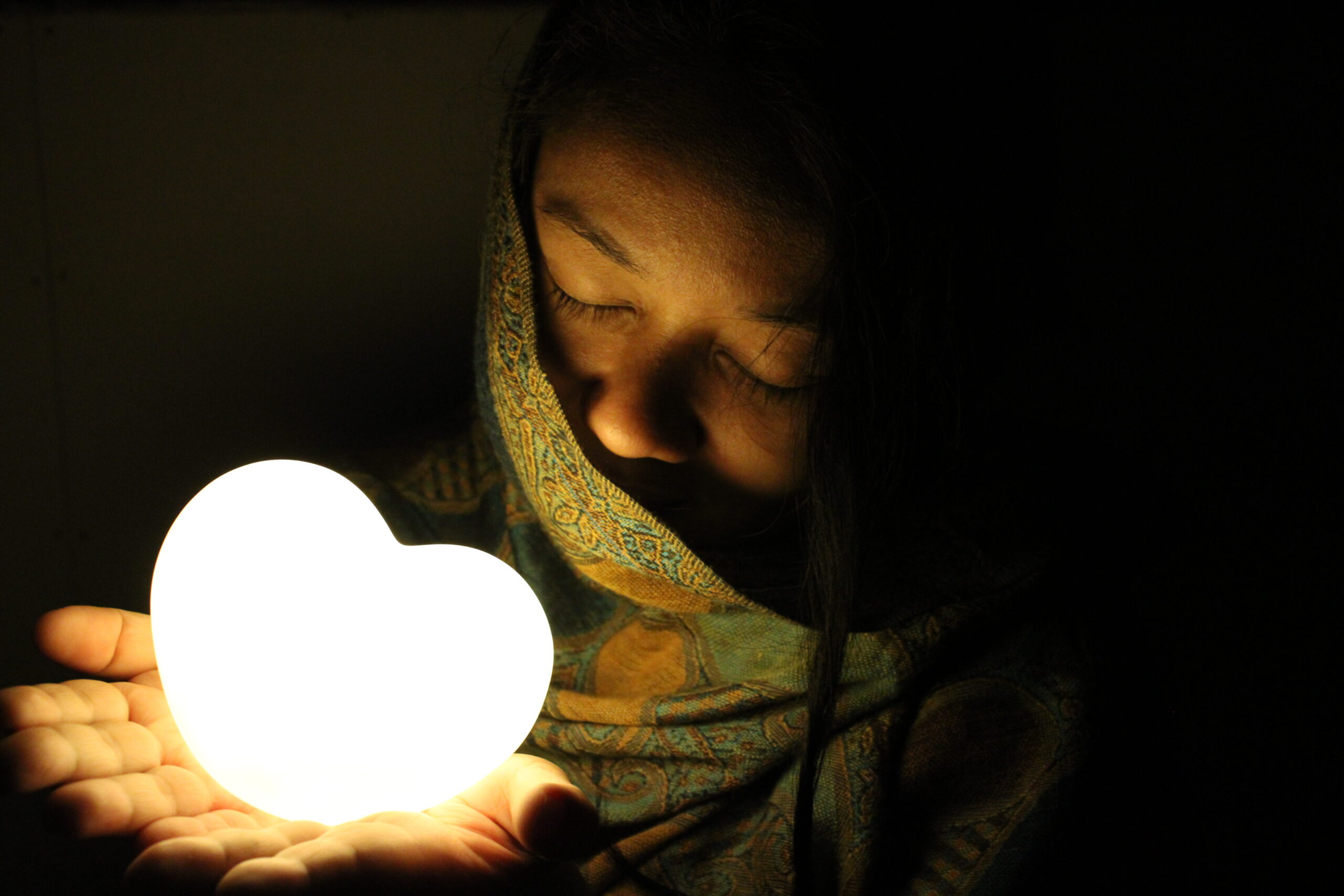 A girl draped in a patterned shawl gently cradles a glowing heart-shaped light, her eyes closed in a moment of tranquility against a dark backdrop.